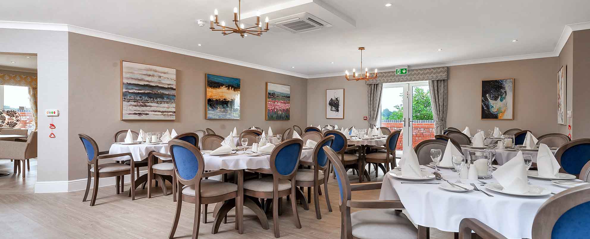 An elegant dining room with multiple set tables, grey and blue upholstered chairs, and paintings on the walls under a coffered ceiling with a chandelier.