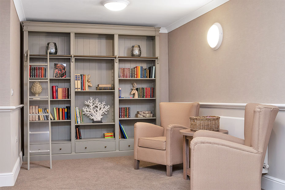 A well-organised library space with a large built-in bookshelf filled with books, decorative items, and storage baskets. Two pink armchairs are placed in front of the bookshelf, and a round wall light is above.