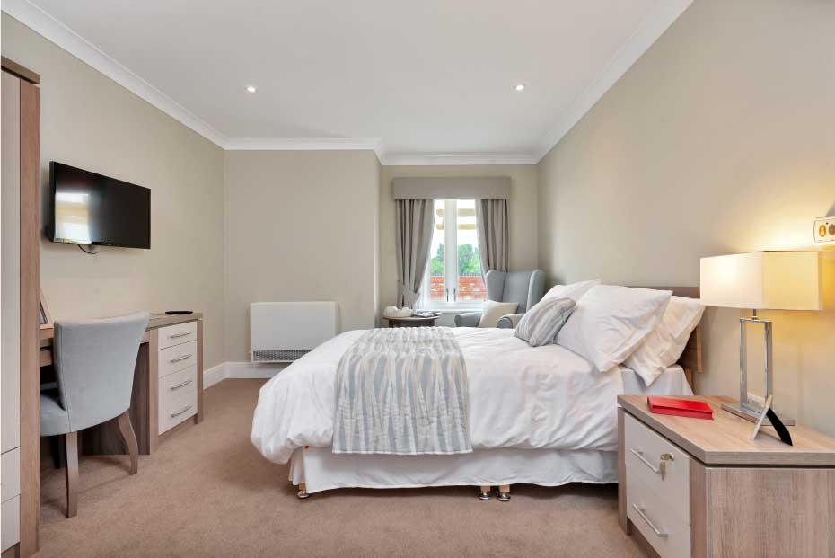 A well-lit, modern bedroom with a large bed, white bedding, a cream throw blanket, two bedside tables with lamps, a desk and chair, and a wall-mounted television.