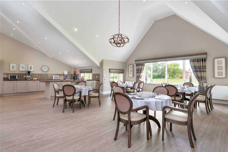 A bright, spacious dining area with round tables, cushioned chairs, a vaulted ceiling, and large windows overlooking greenery. The room has a counter with appliances and is tastefully decorated.