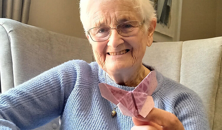 An elderly lady with white hair, wearing a blue  jumper, holds a purple patterned origami butterfly